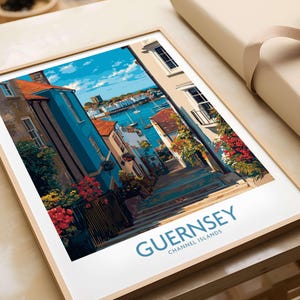 Op de afbeelding: Een ingelijste kunstdruk met een kleurrijke straatscène in Guernsey, Kanaaleilanden. Het kunstwerk toont gebouwen met rode daken, blauwe muren en levendige bloemen. De tekst "GUERNSEY CHANNEL ISLANDS" staat in blauw onderaan.