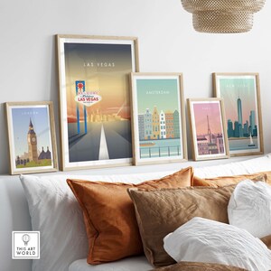 May include: Five framed city skyline prints with a white background. The prints feature the cities of London, Las Vegas, Amsterdam, Paris, and New York City. The prints are arranged in a row on a white wall above a white couch with brown pillows.