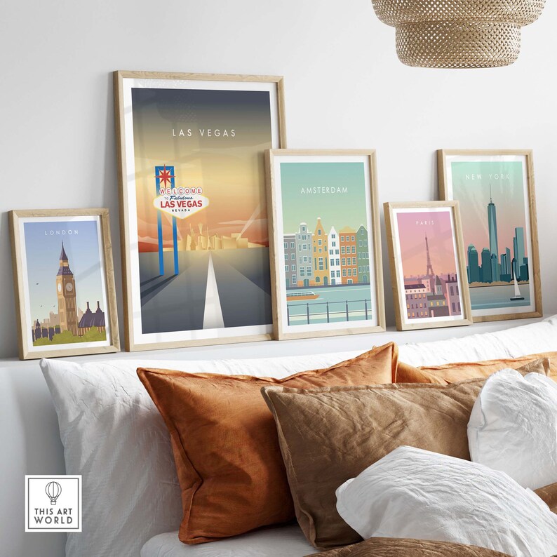 May include: Five framed city skyline prints with a white background. The prints feature the cities of London, Las Vegas, Amsterdam, Paris, and New York City. The prints are arranged in a row on a white wall above a white couch with brown pillows.