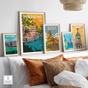 Op de afbeelding: Vijf ingelijste reisposters met illustraties van beroemde bezienswaardigheden. De posters zijn gerangschikt op een witte muur boven een bed met bruine en witte kussens. De posters tonen de volgende steden: Amsterdam, Portofino, Rome, New York en Madrid.