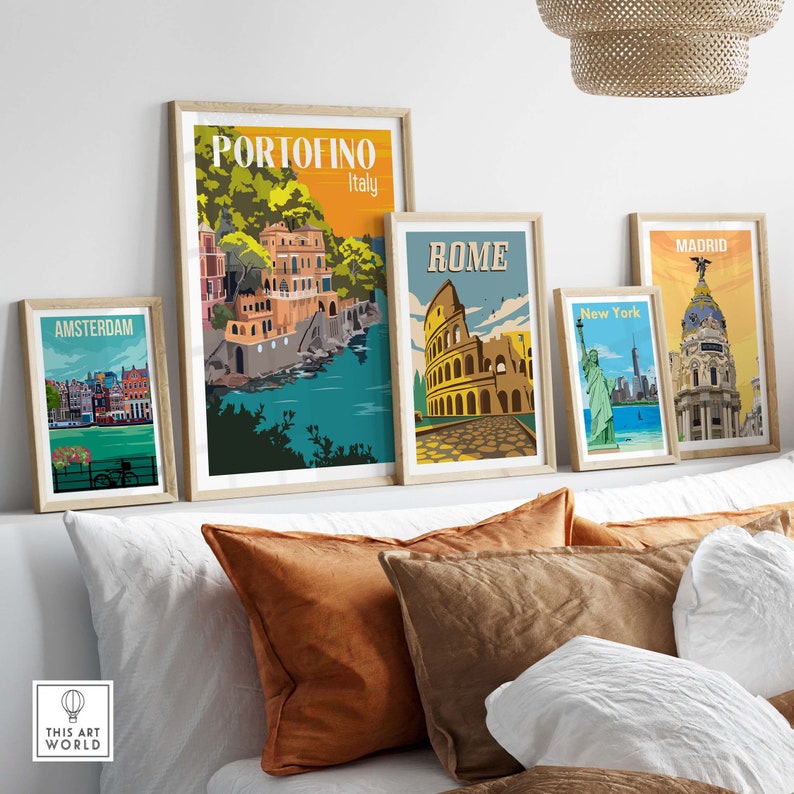 Op de afbeelding: Vijf ingelijste reisposters met illustraties van beroemde bezienswaardigheden. De posters zijn gerangschikt op een witte muur boven een bed met bruine en witte kussens. De posters tonen de volgende steden: Amsterdam, Portofino, Rome, New York en Madrid.