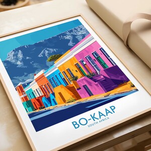 Peut inclure: Une illustration colorée d'une rangée de maisons à Bo-Kaap, en Afrique du Sud, avec la montagne de la Table en arrière-plan. Les maisons sont peintes de couleurs vives, notamment le rose, le jaune, le bleu et le vert. L'illustration est encadrée dans un cadre en bois.