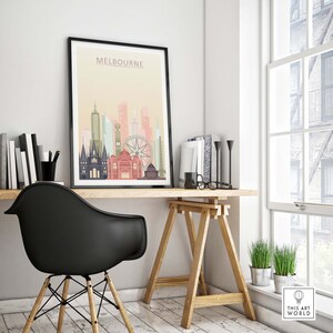 May include: A black framed print with a colorful illustration of the Melbourne skyline. The print is hanging on a white wall above a wooden desk with a black chair and a wooden easel.