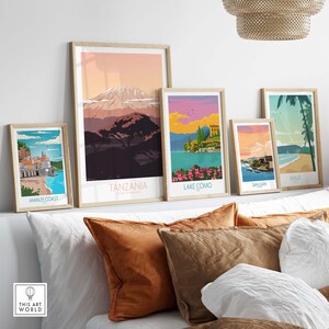 Puede incluir: Cinco p&oacute;sters de viaje enmarcados con ilustraciones coloridas de diferentes destinos. Los p&oacute;sters est&aacute;n dispuestos en una pared blanca sobre un sof&aacute; blanco con almohadas marrones. Los p&oacute;sters presentan los siguientes destinos: Costa Amalfitana, Tanzania, Lago Como, San Juan y Maui.