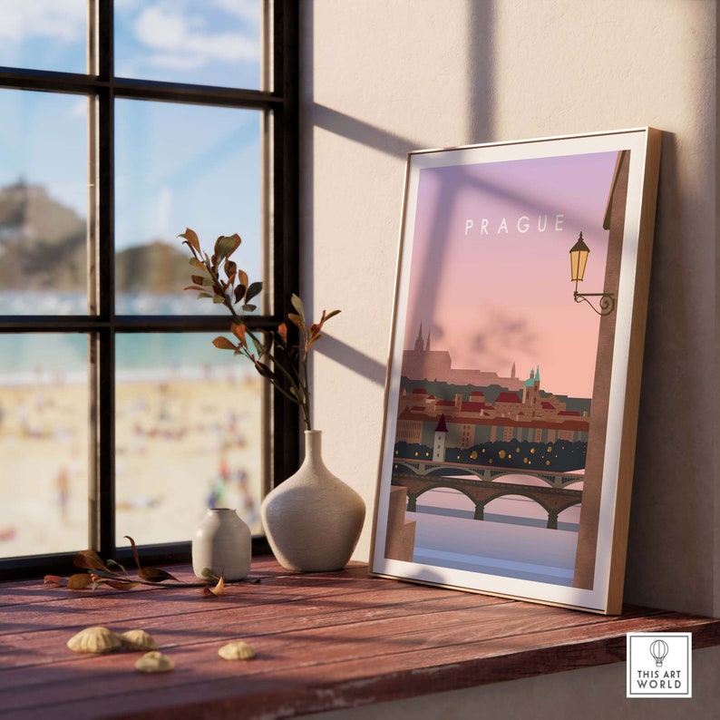 May include: A framed art print featuring a stylized illustration of Prague, with the city's skyline, bridges, and a street lamp. The print is displayed on a wooden surface next to a window with a view of a beach. The word "PRAGUE" is at the top of the print.
