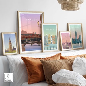 May include: A collection of framed travel art prints featuring iconic cityscapes. The prints showcase Prague, Amsterdam, Paris, New York, and London, each with a unique color palette and design. The frames are light-colored wood, and the prints are displayed on a bed with pillows.