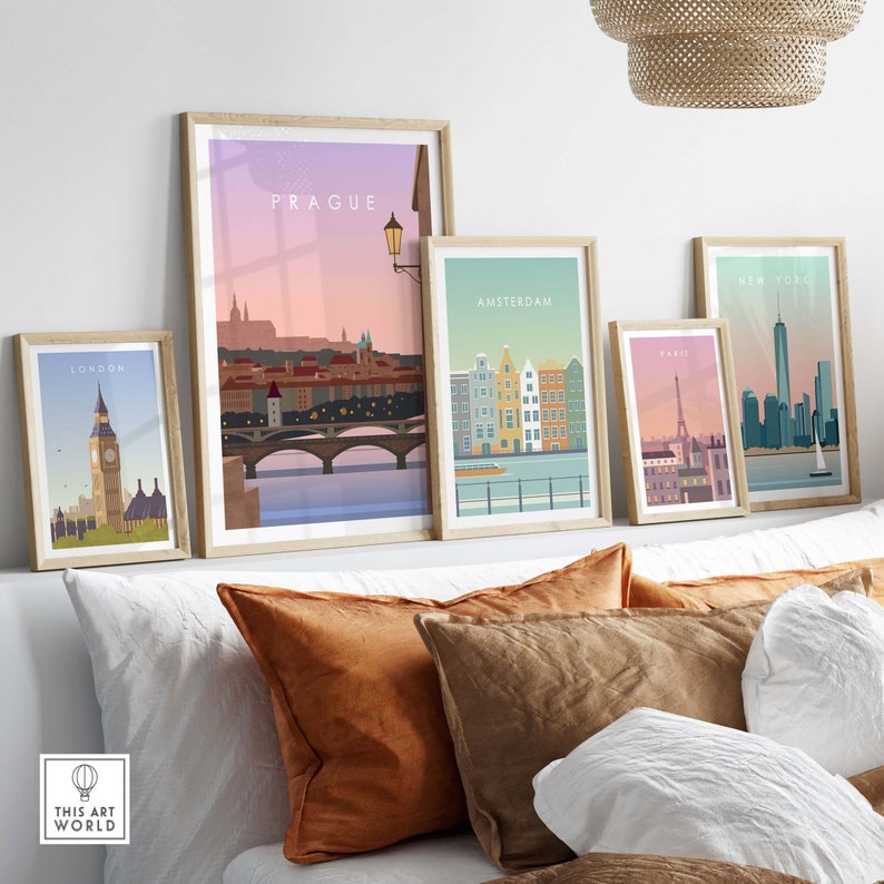 May include: A collection of framed travel art prints featuring iconic cityscapes. The prints showcase Prague, Amsterdam, Paris, New York, and London, each with a unique color palette and design. The frames are light-colored wood, and the prints are displayed on a bed with pillows.