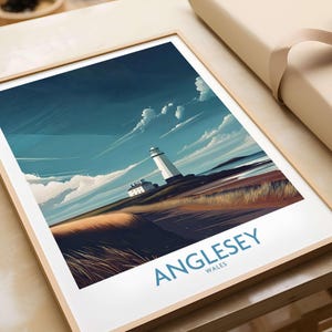 May include: Framed art print featuring a coastal landscape with a white lighthouse under a blue sky with clouds. The print has a minimalist style with the word "Anglesey" in blue text at the bottom. The artwork is in a wooden frame.