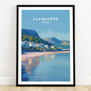 May include: A framed art print featuring a coastal landscape of Llandudno, Wales. The artwork depicts a beach, calm water, and a town nestled at the base of a mountain range under a blue sky. The text "LLANDUDNO Wales" is at the top.