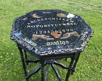 May include: An octagonal black table with a glossy finish featuring a white alphabet, numbers 1-9, and 0, along with celestial sun and moon graphics, the words hello, good bye, Salem, 1692, and two hand illustrations with a heart-shaped planchette.