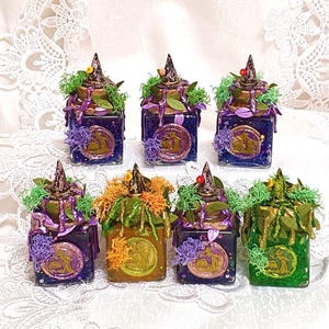 Könnte beinhalten: Sieben dekorative Gläser mit Hexenhutdeckeln, verziert mit Moos, Blättern und einem kreisförmigen Emblem. Die Gläser sind in verschiedenen Farben gehalten, darunter Lila, Grün und Gold. Die Gläser sind auf einer weißen Spitzentischdecke angeordnet.