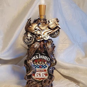 May include: A decorative lamp made from a Jack Daniel's bottle, featuring a motorcycle and Route 66 emblem. The lamp has a gold-colored finish and a faux candle at the top. The base is made of wood.