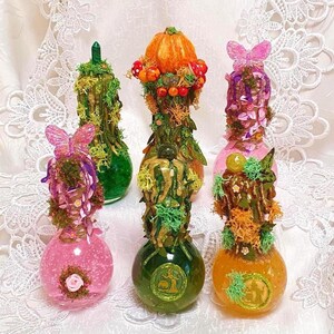 Könnte beinhalten: Eine Sammlung dekorativer Glasflaschen mit aufwendigen, von der Natur inspirierten Designs. Jede Flasche weist eine einzigartige Kombination von Farben auf, darunter Grün, Rosa und Orange, und ist mit Moos, Schmetterlingen und Miniaturkürbissen verziert. Die Flaschen sind auf einer weißen Spitzentischdecke angeordnet.
