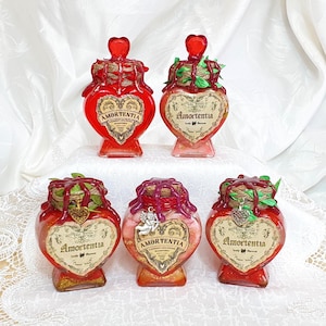 Può includere: Cinque bottiglie di vetro a forma di cuore riempite di liquido rosso e con etichette dorate. Ogni bottiglia ha un tappo di sughero ed è decorata con foglie verdi e spago. Le etichette riportano la scritta "Amortentia" e presentano un motivo a forma di cuore.