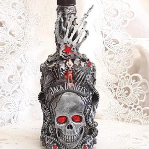 Può includere: Una bottiglia decorativa di Jack Daniel's con un design a teschio. La bottiglia argentata presenta dettagli rossi, un teschio con occhi rossi e un tappo nero. È impreziosita da una mano scheletrica e una stella rossa.
