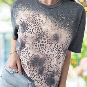 May include: A dark gray t-shirt with a bleached leopard print design. The short-sleeved shirt features a faded, spotted pattern in shades of tan and gray, creating a unique, distressed look. The top is a casual wear item.