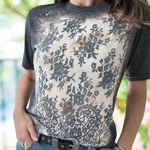 May include: A gray short-sleeved t-shirt with a bleached, distressed effect. The front features a large, intricate floral lace pattern in shades of gray and white. The shirt has a crew neck and a relaxed fit.