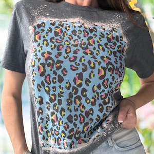 May include: Dark gray t-shirt with a light blue leopard print design. The leopard print features black, pink, and yellow spots. The shirt has a bleached, distressed look around the print.