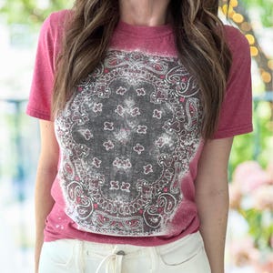 May include: A short-sleeved, faded red t-shirt featuring a distressed bandana print in black, white, and red. The graphic is centered on the front of the shirt. The shirt is paired with white pants.