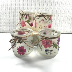 May include: Four small, clear glass jars decorated with pressed flowers in shades of pink, purple, and white. Each jar is wrapped with twine and tied in a bow. The jars are arranged on a small wooden block.
