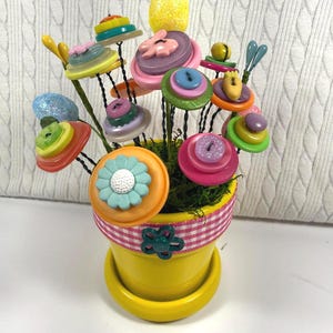 May include: A whimsical arrangement of button flowers in a yellow pot. The flowers are made from colorful buttons and sit atop wire stems. The pot is decorated with a pink and white gingham ribbon and a green flower-shaped embellishment.