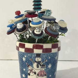 May include: A decorative arrangement featuring multiple wire stems topped with stacked colorful buttons in shades of blue, red, white, and black. The stems are set in a small ceramic pot with a blue snowy background and three snowmen.