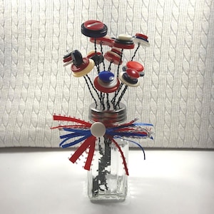 May include: A decorative arrangement of button flowers in a clear glass jar. The button flowers are made of red, white, and blue buttons, and the jar is decorated with red and blue ribbons. The arrangement is a patriotic-themed craft.