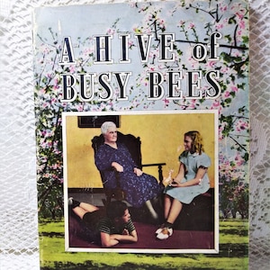 May include: Vintage book cover titled "A Hive of Busy Bees." The cover features a floral background with pink blossoms and the title in bold white letters. A framed image shows three people. The book is displayed on a black stand.