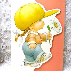 May include: A vintage greeting card featuring a cartoon illustration of a child wearing a yellow cap, orange shirt, and blue pants, holding a daisy. The card has a peach-colored background and a white lace border.