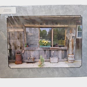 May include: A framed photograph of a rustic wooden structure with a window overlooking a garden. A planter box with yellow flowers sits on the window ledge. The image includes a small wooden box, a barrel, and gardening tools.