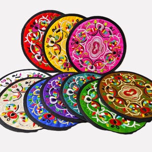 May include: A collection of colorful, embroidered round coasters. The coasters feature intricate floral designs in various colors, including red, yellow, pink, white, purple, and green, with black edging. The coasters are arranged in a fan shape.