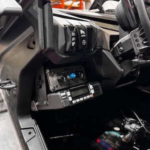 May include: A black and silver radio transceiver with a digital display and a microphone mounted in a vehicle dashboard. The radio has a black and silver control panel with a knob and a blue button. The text "TITOX-115" is visible on the control panel.