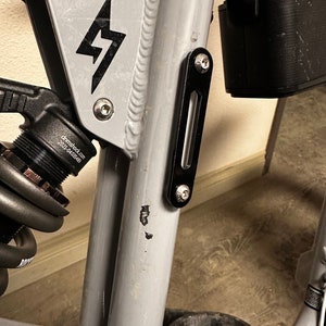 May include: Close-up of a gray bicycle frame with a black lightning bolt logo. The frame has a black metal bracket with a screw on the side. The frame is attached to a black battery pack.