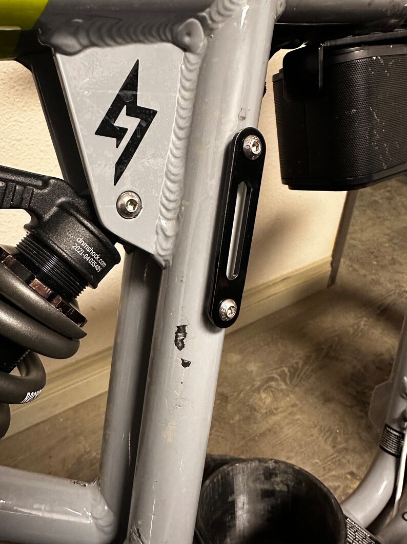 May include: Close-up of a gray bicycle frame with a black lightning bolt logo. The frame has a black metal bracket with a screw on the side. The frame is attached to a black battery pack.