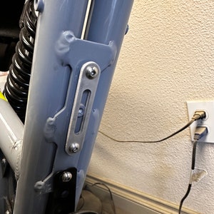 May include: Close-up of a gray metal bike frame with a silver metal bracket attached to it. The bracket has two screws and a slot in the middle. The bike frame is partially visible in the background.