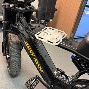 May include: Black Ariel Rider electric bike with a silver rack. The bike has a digital display, black handlebars, and a black seat. The Ariel Rider logo is in yellow. The tires are thick and black.