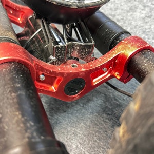 May include: Close-up of a red metal bike frame with a black logo. The frame is attached to two black handlebars with a black and silver suspension system.