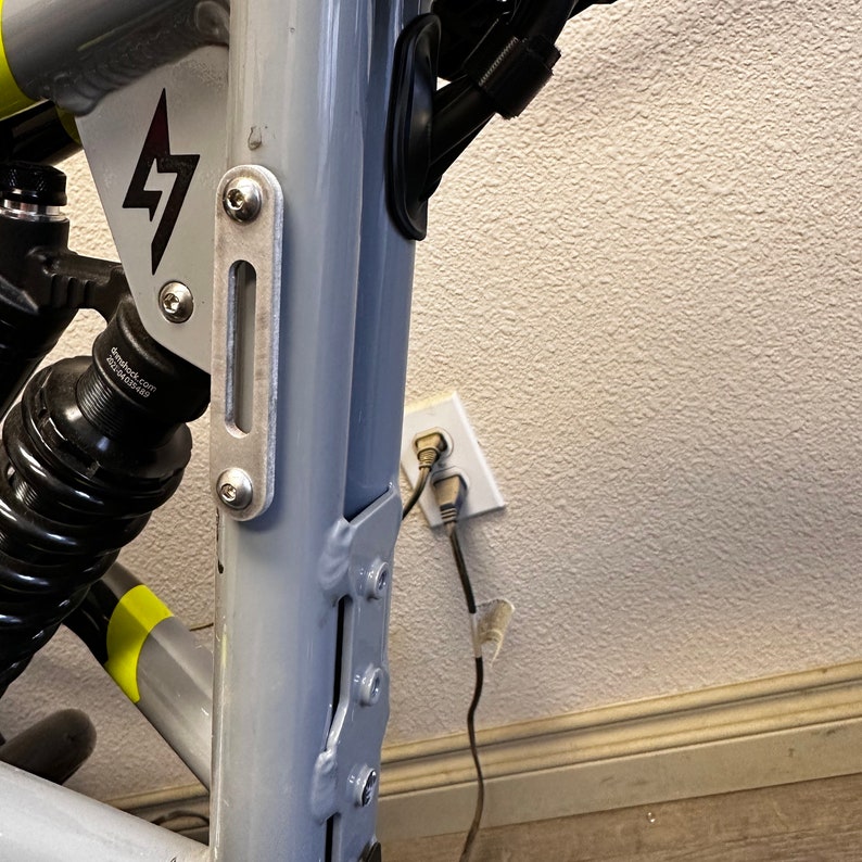 May include: A close-up of a gray electric bike frame with a black shock absorber and a silver metal plate with two screws. The bike is plugged into a wall outlet with a black cord.