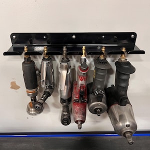Puede incluir: Un soporte de herramientas de metal negro montado en una pared blanca, que sostiene seis herramientas neumáticas diferentes. Las herramientas son de varios tamaños y colores, incluyendo negro, plateado y rojo. El soporte tiene un estante para colgar las herramientas.