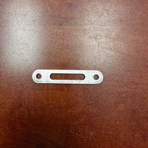 May include: A silver metal strap with two holes and a rectangular cutout in the center.