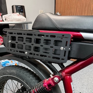May include: A black metal cargo rack attached to a red electric bike. The rack is rectangular with a grid pattern and the text "CUTFAB E". The bike has a black seat and tire, with red and silver accents.