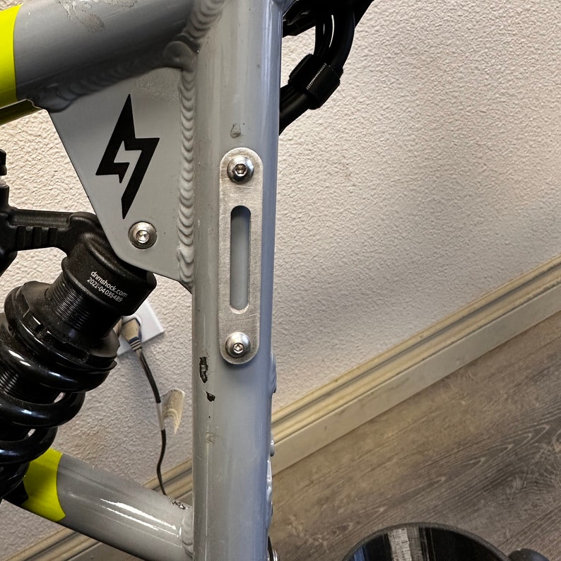 May include: Close-up of a gray bicycle frame with a black lightning bolt logo and a silver metal plate with two screws. The plate has a slot cut into it.