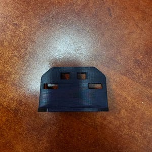 May include: A black, 3D-printed plastic component with a rectangular shape and three rectangular cutouts. The component has a flat base and a slightly curved top edge. The object is on a brown surface.