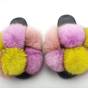 May include: A pair of black fuzzy slides with pink, yellow, and peach colored fur pom-poms.