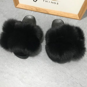 May include: A pair of black faux fur slides. The slides have a black rubber sole and are covered in fluffy black fur.