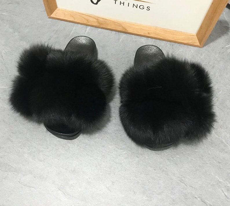 May include: A pair of black faux fur slides. The slides have a black rubber sole and are covered in fluffy black fur.