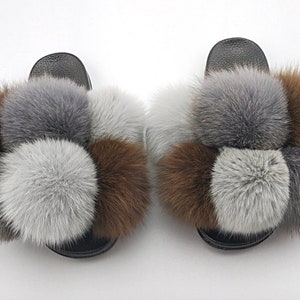 May include: A pair of fuzzy slides with brown, gray, and white pom-poms. The slides have a black base and are made of faux fur.