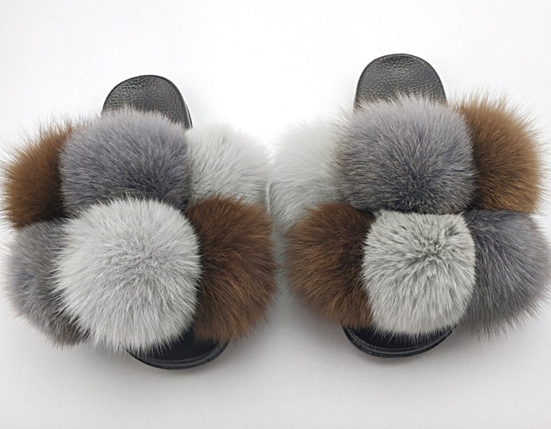 May include: A pair of fuzzy slides with brown, gray, and white pom-poms. The slides have a black base and are made of faux fur.