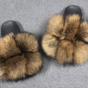 May include: A pair of brown and black fur slides. The slides have a black rubber base and are covered in fluffy brown fur.