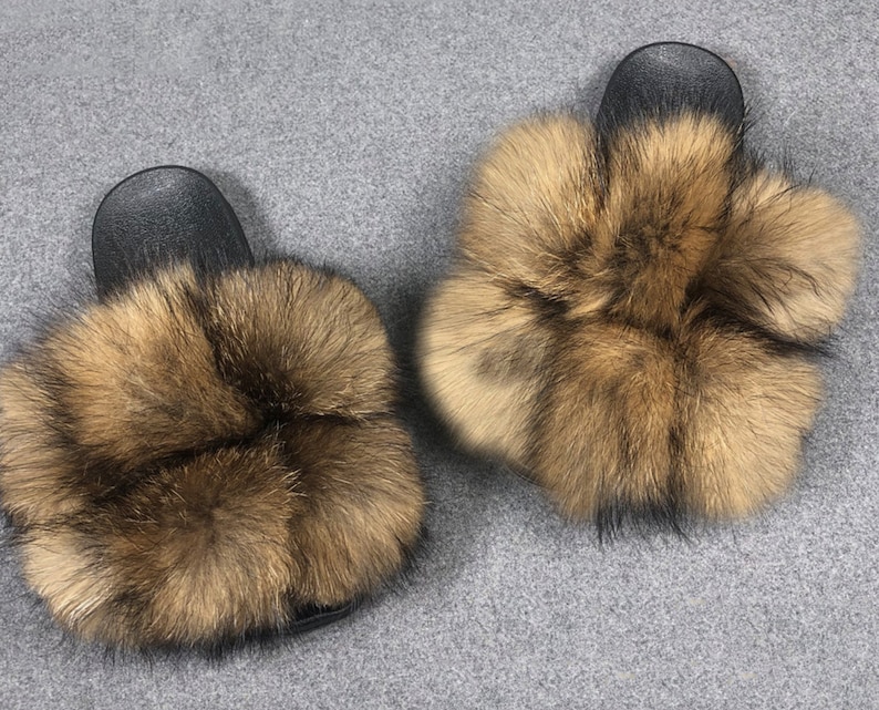 May include: A pair of brown and black fur slides. The slides have a black rubber base and are covered in fluffy brown fur.
